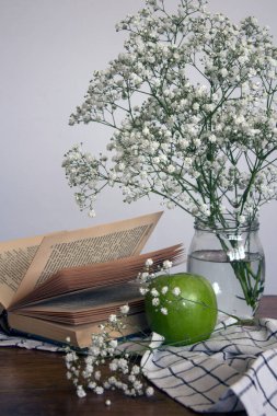 Küçük beyaz çiçekli cam kavanoz, yeşil elma ve açık bir kitap. Basit kompozisyon, beyaz arka planda çingene çiçekleri ile yaşam devam ediyor.. 
