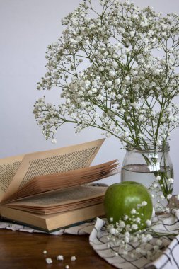 Küçük beyaz çiçekli cam kavanoz, yeşil elma ve açık bir kitap. Basit kompozisyon, beyaz arka planda çingene çiçekleri ile yaşam devam ediyor.. 