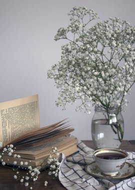 Basit kompozisyon, küçük bir fincan çay, açık kitap ve cam kavanozda çingene çiçekleri ile hayat..  
