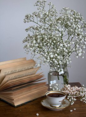 Basit kompozisyon, küçük bir fincan çay, açık kitap ve cam kavanozda çingene çiçekleri ile hayat..  