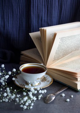 Bir fincan çay ve açık bir kitap. Basit kompozisyon, küçük bir fincan siyah çay, açık kitap ve masanın üzerinde beyaz çigila çiçekleri. Mavi desenli arkaplan. 