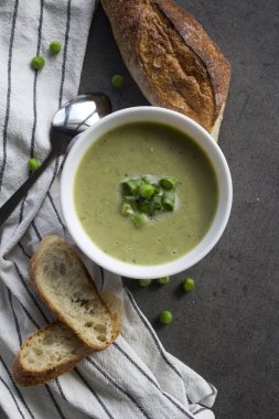 Beyaz kasede yeşil bezelye çorbası, çizgili mutfak havlusunda taze baget ekmeği. Vejetaryen yemeği üst manzara fotoğrafı. Taze konsept yemek.  