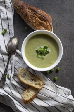 Beyaz kasede yeşil bezelye çorbası, çizgili mutfak havlusunda taze baget ekmeği. Vejetaryen yemeği üst manzara fotoğrafı. Taze konsept yemek.  
