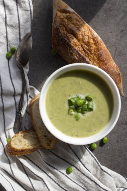 Beyaz kasede yeşil bezelye çorbası, çizgili mutfak havlusunda taze baget ekmeği. Vejetaryen yemeği üst manzara fotoğrafı. Taze konsept yemek.  
