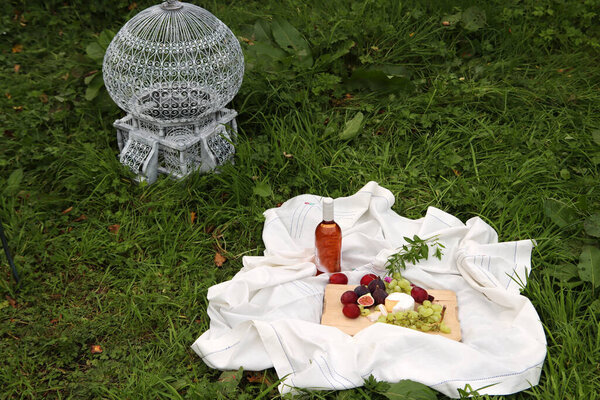 Picnic on the grass. White tablecloth, wooden board with figs, grapes, cheese and plums, bottle of rose wine. Dinner in a garden.