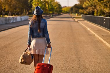 Mavi şapkalı, kot ceketli, etekli, kırmızı valizli, kahverengi çantalı genç bir kadın bir yolculuğa ya da yeni bir hayata başlamak için yolda yürüyor..