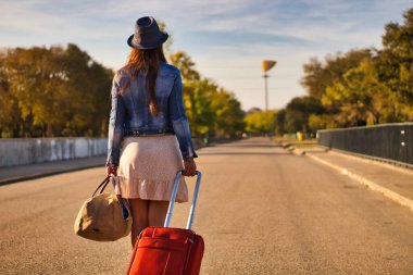 Mavi şapkalı, kot ceketli, etekli, kırmızı valizli ve kahverengi çantalı genç bir kadın bir yolculuğa ya da yeni bir hayata başlamak için yolda yürüyor..