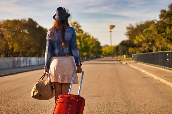 Mavi şapkalı, kot ceketli, etekli, kırmızı valizli ve kahverengi çantalı genç bir kadın bir yolculuğa ya da yeni bir hayata başlamak için yolda yürüyor..