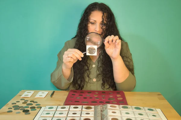 Elinde büyüteçle esmer bir kadın çıplak koleksiyonundaki eski bir madeni paranın detaylarına bakıyor. Tahta bir masada albümü ve açık yeşil bir arka planı var..