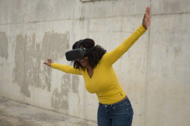 Afro saçlı, sarı tişörtlü, beton duvar arkasında sanal gözlüklerle oynayan Afrikalı Amerikalı bir kadın..