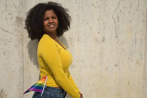 Afro saçlı, sarı tişörtlü ve beton arka planda eşcinsel gurur bayrağı taşıyan Afro-Amerikalı bir kadın..