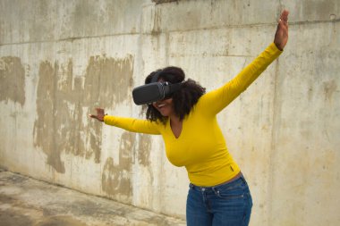 Afro saçlı, sarı tişörtlü, beton duvar arkasında sanal gözlüklerle oynayan Afrikalı Amerikalı bir kadın..
