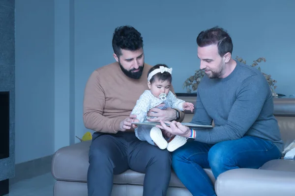 Eşcinsel evli çift evdeki kanepede kızlarına masal okuyor. Aile çeşitliliği.