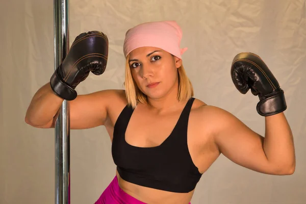 Female pole dancer with pink cancer scarf and boxing gloves holding on to the bar.