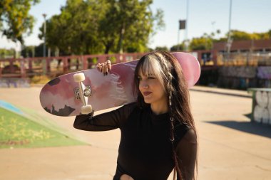 Punk tarzı ve heterokromisi olan hoş bir genç kız kaykay parkında düşünceli bir bakışla kaykayı omzunun üstünde tutuyor..