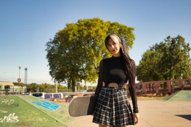 Punk tarzı ve heterokromisi olan genç ve güzel bir kız elinde kaykayla kaykay parkındaki kameraya bakıyor..