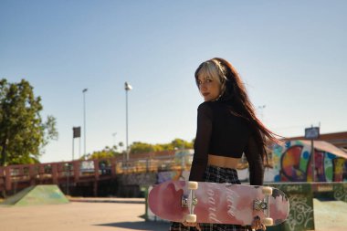 Punk tarzı ve heterokromisi olan hoş bir genç kız kaykay parkında elleri arkada kaykay tutuyor..