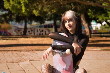 Genç ve güzel bir kızın portresi heterokromi ve punk stili kaldırımda oturmuş kaykayına sarılıyor..