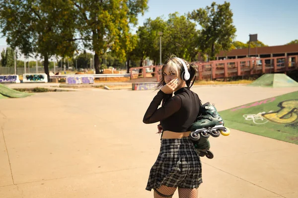 Young girl with pigtails, punk style, white headphones and inline skates hanging from the shoulder on their backs with one hand on their face looking at the camera.