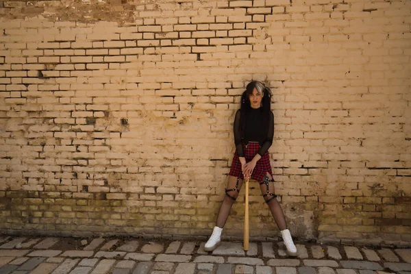Saçları örgülü ve punk stili genç ve güzel bir kız. Arka planda sarı tuğla bir duvarla bir beyzbol sopasına yaslanıyor..