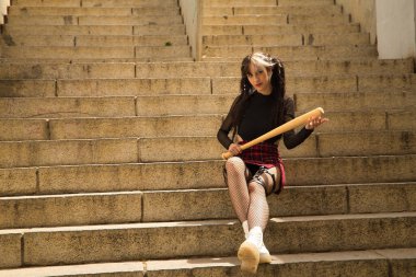 Genç ve güzel bir kız, saçları örgülü, heterokromi ve punk stili merdivenlerde elinde beyzbol sopasıyla oturuyor..