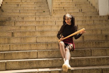 Young and beautiful girl with pigtails, heterochromia and punk style sitting on some stairs holding a baseball bat in her hands. clipart