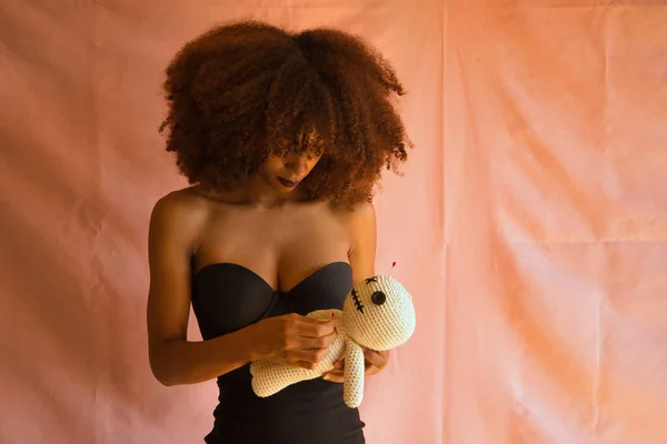 Siyah elbiseli genç Latin bir kadın elinde tuttuğu bir vudu bebeğinin kalbine pembe arka planda iğne batırıyor..