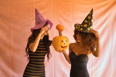 Cadı şapkalı genç İspanyol ve Latin kadınlar pembe arka planda bir Cadılar Bayramı balkabağını ortaya çıkarıyorlar..