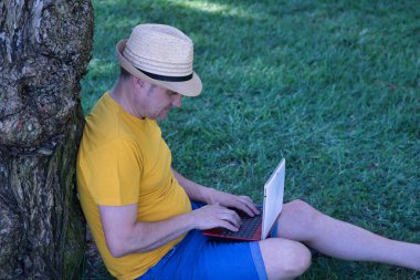 İspanyol yetişkin bir adam parkta çimlerde oturur, dizüstü bilgisayarıyla çalışırken bir ağaca yaslanır..