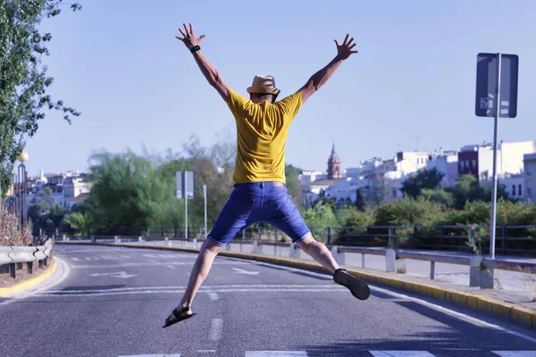 Şapkalı, hardal renkli tişörtlü ve kısa kotlu yetişkin İspanyol bir adam şehrin ortasında yaya geçidinden atlıyor..