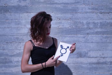 Genç yetişkin İspanyol transseksüel kız, gri çimento arka planında, bir not defterinde cinsiyet sembolünü işaret ediyor. Transseksüel konsept, kapsama ve çeşitlilik.