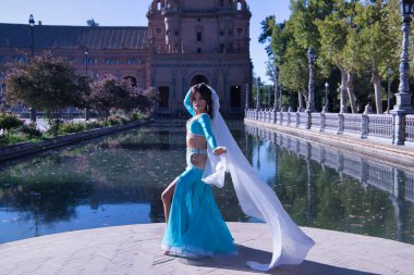 Orta yaşlı, turkuaz elbiseli, yapay elmaslı İspanyol kadın, beyaz peçeli göbek dansı. Göbek dansı konsepti.