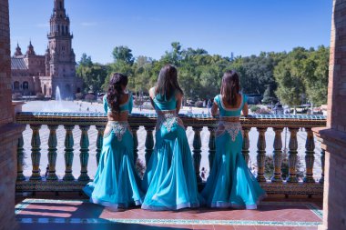 Üç orta yaşlı İspanyol kadın, turkuaz elbiseler ve yapay elmaslar giyiyorlar, göbek dansında, fayanslı korkulukların yanında poz veriyorlar. Göbek dansı konsepti.