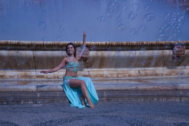 Orta yaşlı İspanyol kadın, su yeşili bir elbise giyiyor, yapay elmasları var, göbek dansı için, etrafında sabun köpükleriyle çeşmede oturuyor. Göbek dansı konsepti.