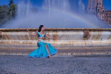 Orta yaşlı İspanyol kadın, turkuaz kostümlü ve yapay elmaslı, göbek dansı yapan, gökkuşağının oluştuğu bir çeşmede oturan. Göbek dansı konsepti.