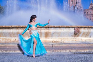 Orta yaşlı İspanyol kadın, turkuaz kostümlü ve yapay elmaslı, göbek dansı için, bir gökkuşağının oluştuğu bir çeşmenin önünde dans ediyor. Göbek dansı konsepti.