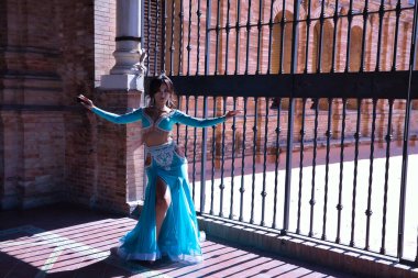 Orta yaşlı İspanyol bir kadın, yapay elmaslı turkuaz bir elbise giyiyor, göbek dansı için, demir ızgaranın yanında dans ederken figürler yapıyor, arka plan ışığı. Göbek dansı konsepti.