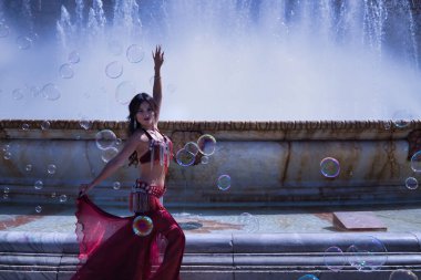 Orta yaşlı İspanyol bir kadın, suni elmaslı kırmızı bir elbise giyiyor, göbek dansı için, etrafında sabun köpükleriyle çeşmenin önünde dans ediyor. Göbek dansı konsepti.