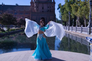 Orta yaşlı, turkuaz elbiseli, yapay elmaslı İspanyol kadın, beyaz peçeli göbek dansı. Göbek dansı konsepti.