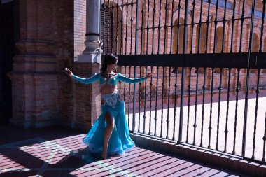 Orta yaşlı İspanyol bir kadın, yapay elmaslı turkuaz bir elbise giyiyor, göbek dansı için, demir ızgaranın yanında dans ederken figürler yapıyor, arka plan ışığı. Göbek dansı konsepti.