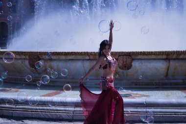 Orta yaşlı İspanyol bir kadın, suni elmaslı kırmızı bir elbise giyiyor, göbek dansı için, etrafında sabun köpükleriyle çeşmenin önünde dans ediyor. Göbek dansı konsepti.