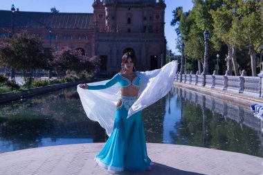 Orta yaşlı, turkuaz elbiseli, yapay elmaslı İspanyol kadın, beyaz peçeli göbek dansı. Göbek dansı konsepti.