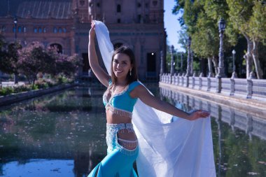 Orta yaşlı, turkuaz elbiseli, yapay elmaslı İspanyol kadın, beyaz peçeli göbek dansı. Göbek dansı konsepti.