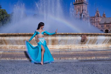 Orta yaşlı İspanyol kadın, turkuaz kostümlü ve yapay elmaslı, göbek dansı için, bir gökkuşağının oluştuğu bir çeşmenin önünde dans ediyor. Göbek dansı konsepti.