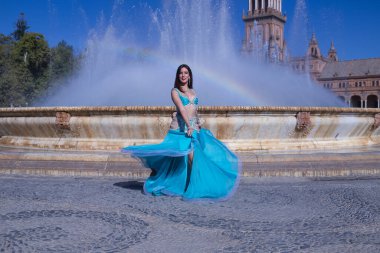 Orta yaşlı İspanyol kadın, turkuaz kostümlü ve yapay elmaslı, göbek dansı için, bir gökkuşağının oluştuğu bir çeşmenin önünde dans ediyor. Göbek dansı konsepti.