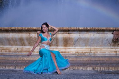 Orta yaşlı İspanyol kadın, turkuaz kostümlü ve yapay elmaslı, göbek dansı yapan, gökkuşağının oluştuğu bir çeşmede oturan. Göbek dansı konsepti.