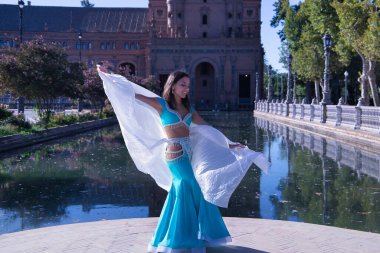 Orta yaşlı, turkuaz elbiseli, yapay elmaslı İspanyol kadın, beyaz peçeli göbek dansı. Göbek dansı konsepti.