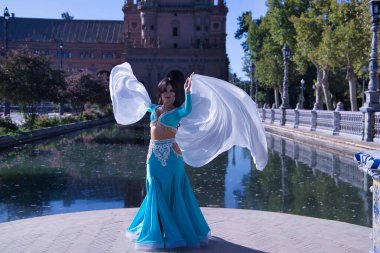 Orta yaşlı, turkuaz elbiseli, yapay elmaslı İspanyol kadın, beyaz peçeli göbek dansı. Göbek dansı konsepti.