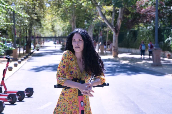 Middle-aged adult Hispanic woman with black curly hair, wearing a colorful outfit, leaning on an electric scooter. Concept scooter, urban mobility, electricity, battery, ecology.