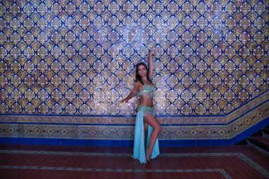 Orta yaşlı, açık yeşil elbiseli, göbek dansı için yapay elmasları olan İspanyol bir kadın. Arka planda güzel fayanslardan oluşan bir duvarla dans ederken figürler yapıyor. Göbek dansı konsepti.